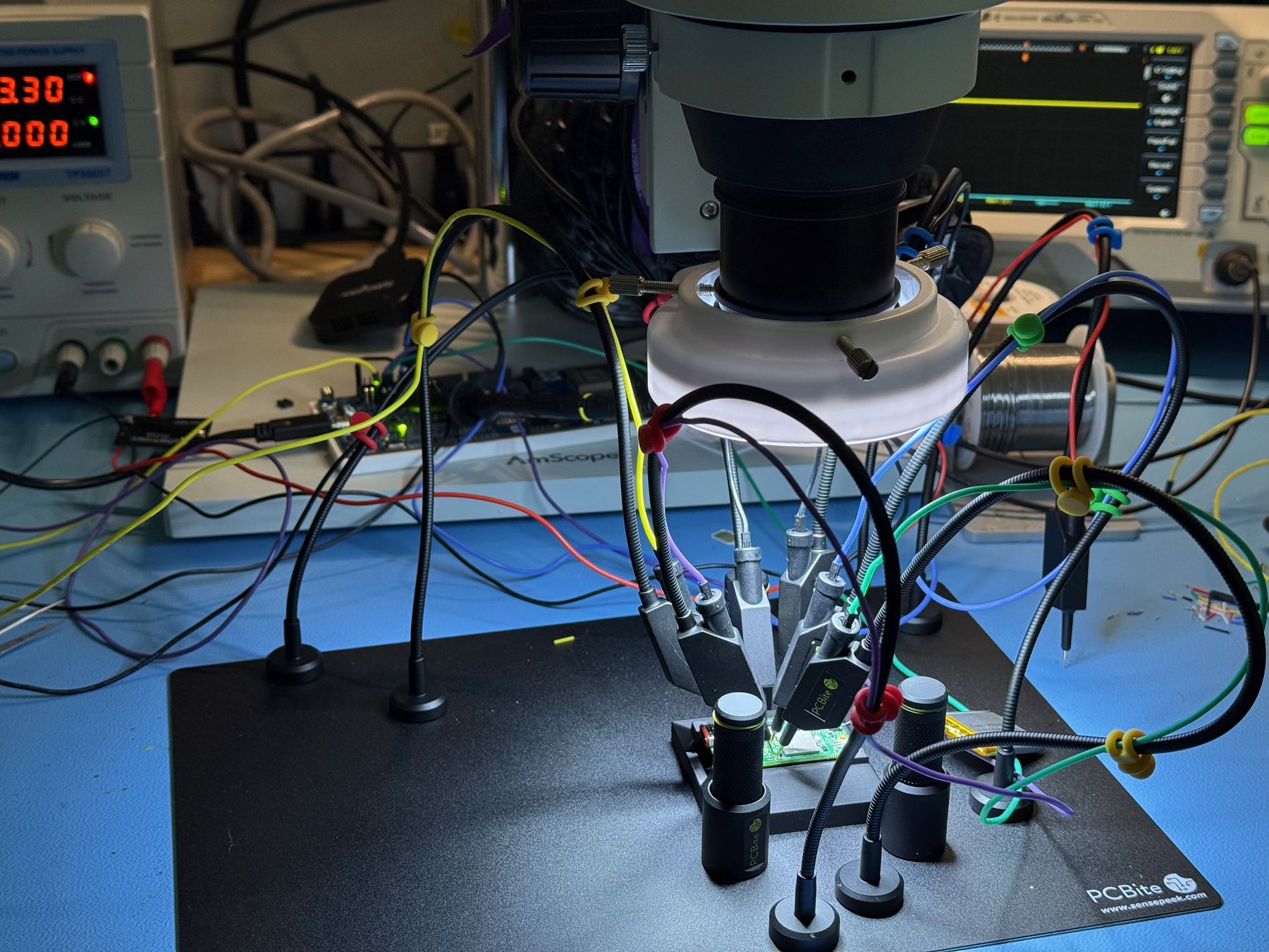 AirGuard board under a microscope on the bench, probed with PCBite fixtures alongside an oscilloscope and bench supply
